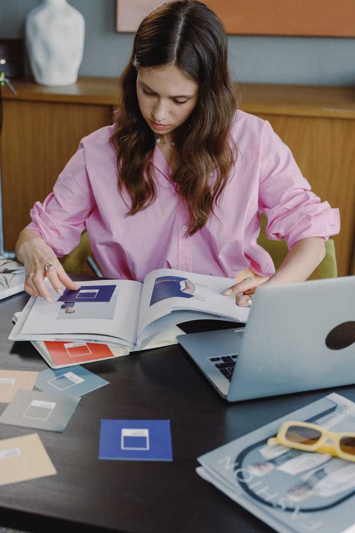 Focused woman in pink blouse flipping through design references at a desk with a laptop.