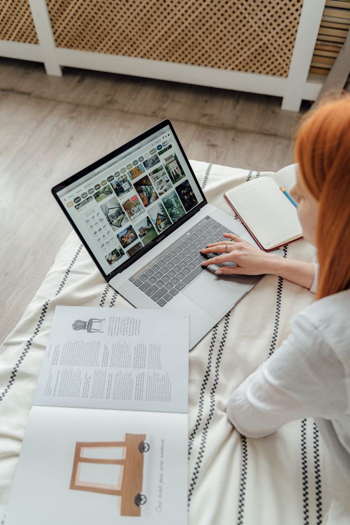 A woman comfortably works on a laptop in a cozy bedroom, surrounded by books and notes.